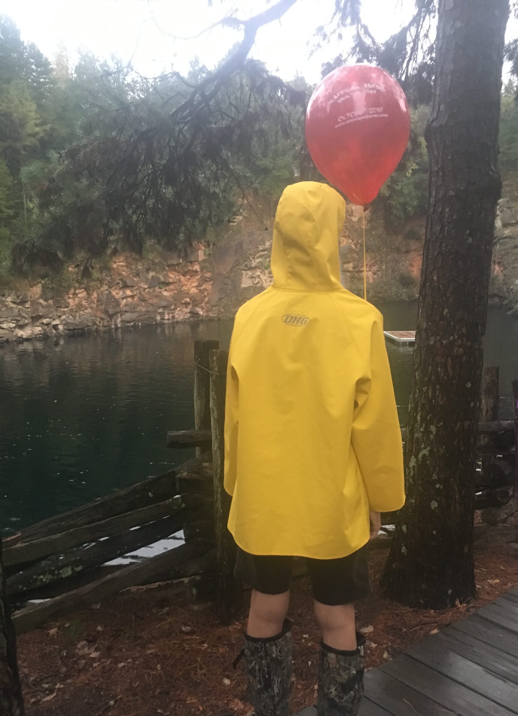 A young man reminscent of Georgie from the movie IT stands with a red balloon wearing a yellow rain coat at the quarry at Carrigan Farms in Mooresville, NC.